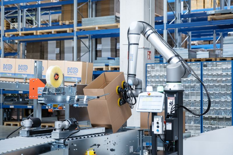 Cobot fixiert Karton in Bodenklappenfalter Cobot fixiert Karton in Bodenklappenfalter